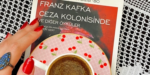 photo from above: a cup with coffee placed on a Kafka book placed on a surface with a tablecloth, next to it a vase with a flower, author: Gizem Nur